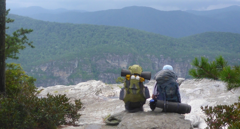Two people wearing backpacks sit on a rock, facing away from the camera. They are looking out over a vast and green mountainous landscape.
